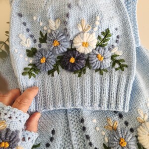 May include: A light blue knitted hat and matching sweater set. The set features embroidered flowers in shades of blue, white, and yellow, with green leaves. The hat has a brim and the sweater has a button-up front. The set is handmade.