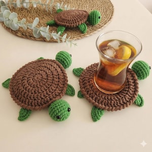 May include: Crocheted turtle coasters in brown and green. The coasters have a round, textured shell and green head and flippers. One coaster holds a glass of iced tea with a lemon slice. A similar turtle coaster sits on a woven mat.