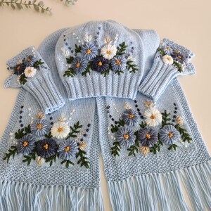 May include: A light blue knitted winter set featuring a hat, scarf, and fingerless gloves, all adorned with embroidered floral designs in shades of blue, white, and gold. The scarf has a fringed edge. The set is designed for cold weather.