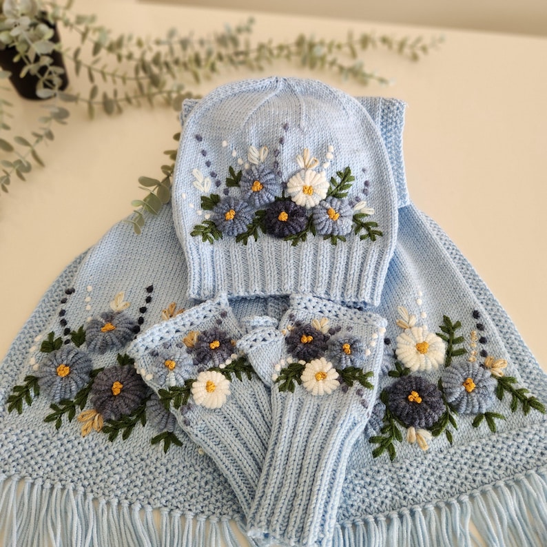 May include: A light blue knitted winter set featuring a hat, scarf, and fingerless gloves, all adorned with embroidered floral designs in shades of blue, white, and yellow. The scarf has a fringe.