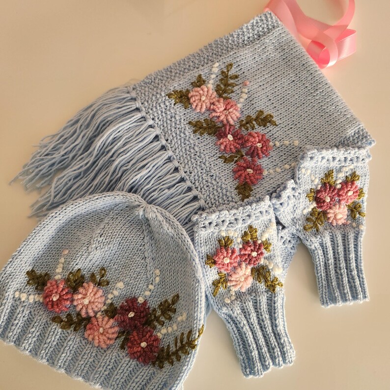 May include: A light blue knitted hat, scarf with fringe, and fingerless gloves set. Each piece is adorned with embroidered pink and burgundy flowers, green leaves, and white beaded accents. A pink ribbon is attached to the scarf.