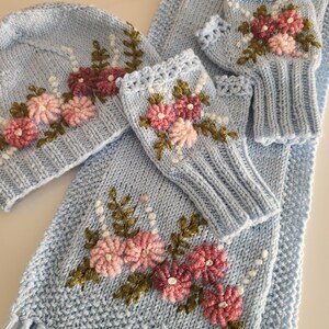 May include: A light blue knitted winter set featuring a hat, scarf, and fingerless gloves. The set is adorned with embroidered pink and burgundy flowers with green leaves and white bead accents. The scarf has a fringed edge.
