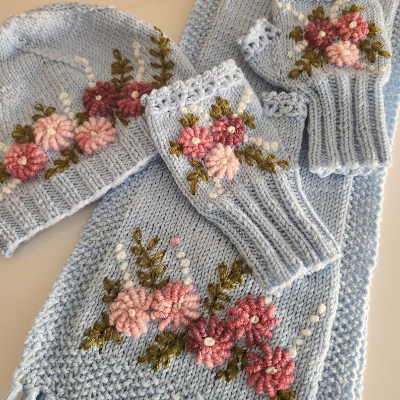 May include: A light blue knitted winter set featuring a hat, scarf, and fingerless gloves. The set is adorned with embroidered pink and burgundy flowers with green leaves and white bead accents. The scarf has a fringed edge.