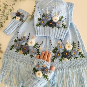 May include: A light blue knitted winter set featuring a hat, scarf, and fingerless gloves, all adorned with embroidered floral designs in shades of blue, white, and yellow. The scarf has a fringed edge.