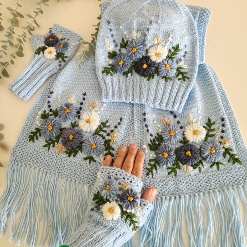 May include: A light blue knitted winter set featuring a hat, scarf, and fingerless gloves, all adorned with embroidered floral designs in shades of blue, white, and yellow. The scarf has a fringed edge.