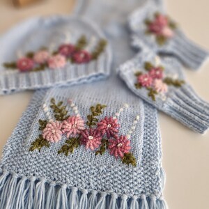 May include: A light blue knitted set featuring a hat, scarf, and fingerless gloves, all adorned with embroidered pink and purple flowers and green leaves. The scarf has a fringed edge. The set is made of soft, textured yarn.