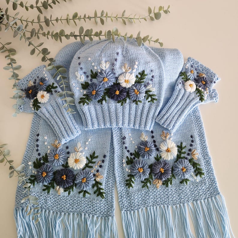 May include: A light blue knitted winter set featuring a hat, scarf, and fingerless gloves, all adorned with embroidered floral designs in shades of blue, white, and gold. The scarf has a fringed edge.