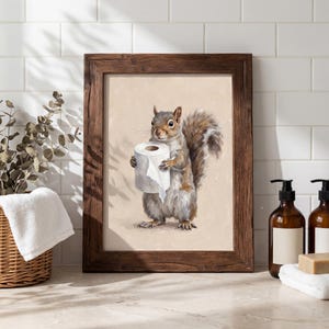 May include: A framed print featuring a squirrel holding a roll of toilet paper. The artwork has a neutral background and is set in a dark brown wooden frame. The squirrel is depicted in shades of gray and brown.
