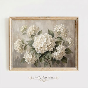 Op de afbeelding: Een ingelijste bloemenprint met een gedetailleerd schilderij van witte hortensia's met groene bladeren tegen een gedempte grijze achtergrond. Het kunstwerk is in een goudkleurige lijst geplaatst, wat een vintage esthetiek creëert. De print is gesigneerd "Artly Nest Prints".