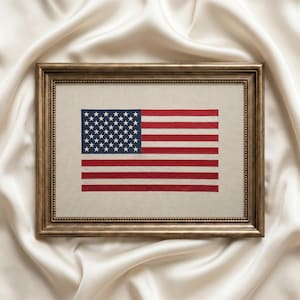 May include: Framed embroidered American flag with red and white stripes and a blue field of white stars. The flag is displayed in a gold-colored frame, set against a backdrop of draped, off-white fabric.