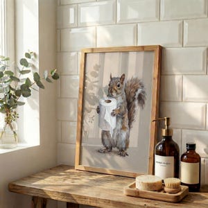 May include: A framed print featuring a watercolor illustration of a squirrel holding a roll of toilet paper. The artwork has a neutral color palette with a wooden frame, displayed on a wooden surface near bathroom accessories.