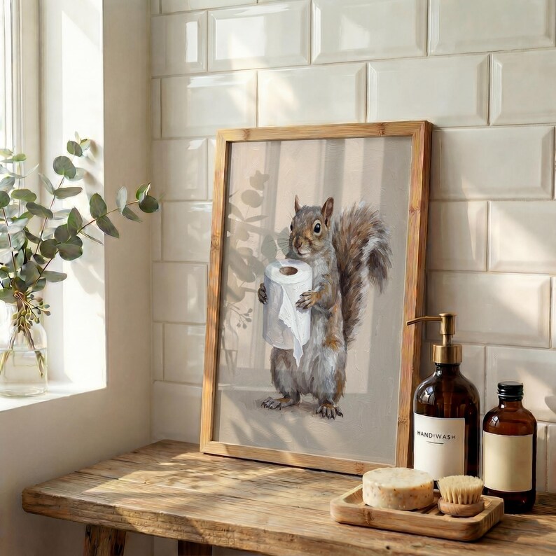 May include: A framed print featuring a watercolor illustration of a squirrel holding a roll of toilet paper. The artwork has a neutral color palette with a wooden frame, displayed on a wooden surface near bathroom accessories.