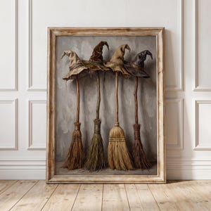 May include: A framed wall art print depicting four witch hats resting atop four wooden broomsticks. The hats are rendered in shades of beige, olive green, tan, and dark brown, while the broomsticks feature natural straw bristles.