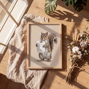 May include: A framed print featuring a detailed illustration of a squirrel holding a roll of toilet paper. The squirrel is depicted in shades of gray and brown, set against a neutral background. The frame is made of light-colored wood.