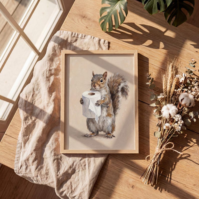 May include: A framed print featuring a detailed illustration of a squirrel holding a roll of toilet paper. The squirrel is depicted in shades of gray and brown, set against a neutral background. The frame is made of light-colored wood.