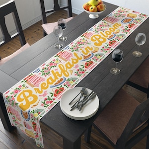 May include: A white table runner featuring a repeating pattern of colorful flowers, strawberries, and breakfast items like toast and croissants. The text "Breakfast in Bloom" is printed in large yellow script across the center of the runner.