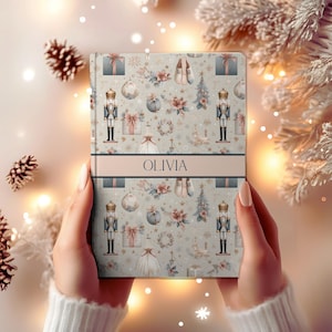 Puede incluir: Un cuaderno personalizado con un diseño navideño festivo. El diseño incluye cascanueces, cajas de regalo, adornos y elementos florales en suaves colores pastel. El nombre "OLIVIA" se muestra en una banda horizontal en la portada.