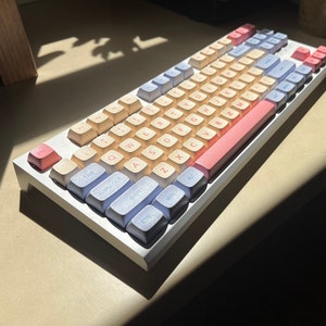 May include: A white mechanical keyboard with pastel blue, pink, and beige keycaps. The keyboard has a pink space bar and a pink escape key.