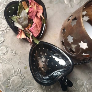 May include: A heart-shaped, black metal tea infuser filled with dried rose petals and leaves. The infuser is open, revealing the contents. A bronze-colored candle holder with star cutouts is also visible.