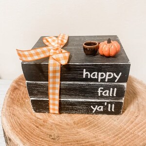 Puede incluir: Un juego de bloques de madera negros decorativos con las palabras "happy fall ya'll" en blanco. Cuenta con una pequeña calabaza naranja, un pequeño cuenco marrón y una cinta de cuadros naranja y blanca. Se muestra sobre una rodaja de madera.