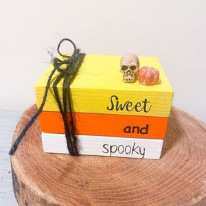 Puede incluir: Tres bloques de madera apilados pintados en amarillo, naranja y blanco con el texto "Sweet and spooky" en ellos. Un pequeño cráneo blanco y una pequeña calabaza naranja están encima de los bloques.