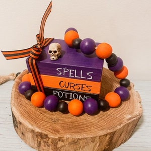 Puede incluir: Tres bloques de madera apilados uno encima del otro con las palabras "Spells", "Curses" y "Potions" escritas en ellos. Una guirnalda de cuentas negras y naranjas está envuelta alrededor de los bloques y un pequeño amuleto de calavera blanca está en el bloque superior.