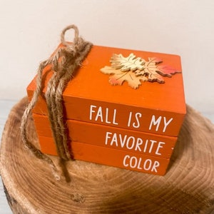 Puede incluir: Una pila de tres bloques de madera naranja con el texto "El otoño es mi color favorito" en blanco. El bloque superior tiene un recorte de hoja de madera.
