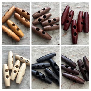 Könnte beinhalten: Eine Auswahl an hölzernen Knebelknöpfen in verschiedenen Brauntönen, von hellem Beige bis zu dunklem Mahagoni. Jeder Knopf hat eine zylindrische Form mit drei Löchern zum Annähen. Die Knöpfe liegen auf einer hellen Holzoberfläche.