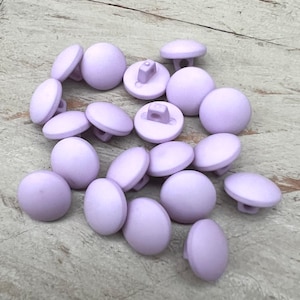 Könnte beinhalten: Eine Ansammlung runder, hellvioletter Knöpfe mit mattem Finish. Die Knöpfe haben eine leicht gewölbte Oberseite und einen Schaft auf der Rückseite zum Annähen. Die Knöpfe liegen auf einer hellen Holzoberfläche.