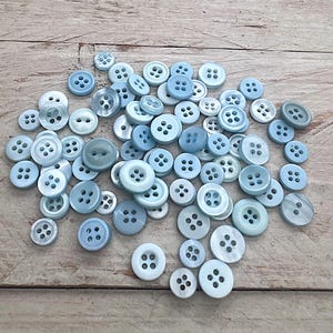 Könnte beinhalten: Eine Ansammlung hellblauer Knöpfe in verschiedenen Größen, auf einer hellen Holzoberfläche verstreut. Die Knöpfe haben vier Löcher und ein leicht schimmerndes Finish, das einen dezenten Glanz erzeugt. Ideal für Näh- und Bastelprojekte.