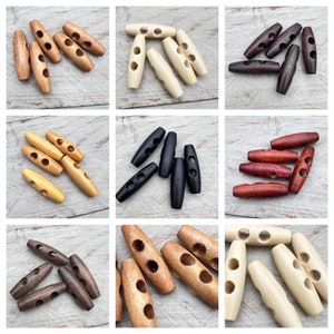 Könnte beinhalten: Sortiment aus Holz-Toggle-Knöpfen in verschiedenen Farben, darunter Naturholz, Braun, Schwarz und Rot. Die Knöpfe haben zwei Löcher und sind oval geformt.