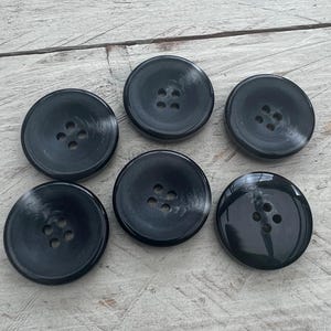 Könnte beinhalten: Sechs schwarze Plastikknöpfe mit jeweils vier Löchern. Die Knöpfe sind rund und haben eine glatte, glänzende Oberfläche.