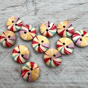 Könnte beinhalten: Eine Sammlung bunter Holzknöpfe mit einem Spiraldesign. Jeder Knopf hat eine Mischung aus roten, gelben, lila und türkisfarbenen Streifen, die von der Mitte ausgehen. Die Knöpfe haben zwei Löcher zum Nähen und sind auf einer hellgrauen Oberfläche angeordnet.
