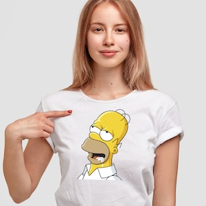 Puede incluir: Una camiseta blanca de manga corta con un estampado gráfico del personaje de dibujos animados amarillo Homer Simpson. El personaje aparece con la zona de la boca bronceada, los ojos muy abiertos mirando hacia arriba y saliva blanca.
