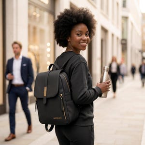 Puede incluir: Una mochila negra con cierre de solapa, herrajes en tono dorado y el texto GASTON LUCA impreso en dorado en la parte frontal. La mochila cuenta con un asa superior, correas ajustables y bolsillos laterales, junto a una botella de agua plateada.
