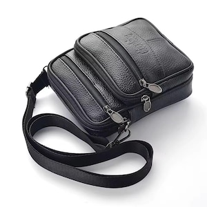 Könnte beinhalten: Schwarze Leder-Umhängetasche mit mehreren Reißverschlussfächern. Die Tasche hat eine strukturierte Oberfläche und einen langen, verstellbaren Riemen. Auf der Oberseite ist das Wort "Wandaiiu" eingeprägt. Für den Transport von Essentials.