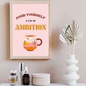 May include: A pink framed print with the text "Pour Yourself A Cup Of Ambition" in orange and brown lettering. A cartoon mug with a desert scene inside is pictured below the text.