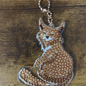 Puede incluir: Un llavero transparente con forma de zorro adornado con numerosos diamantes de imitación de color ámbar. El zorro tiene diamantes de imitación de color rojo oscuro para los ojos y la nariz. Una cadena de bolas y un cierre dorados están unidos a la parte superior del llavero.