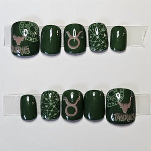 May include: A set of dark green press-on nails. Some nails feature a pink Taurus symbol, a sun design, and the word "TAURUS". Other nails have a geometric pattern. The nails are arranged on a clear strip.