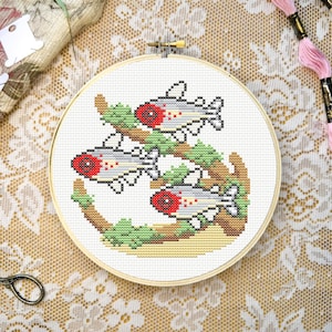 May include: A circular wooden embroidery hoop containing a cross-stitch pattern of three fish with red heads and grey bodies swimming around a brown branch with green moss. The design is stitched on white fabric, centred within the round frame.