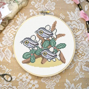 May include: A circular wooden embroidery hoop containing a cross-stitch pattern of three small, light purple fish with large yellow eyes, positioned on a brown branch with green leaves and a yellow sandy base against a white fabric background.