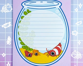 May include: A pink notepad with a blue and white illustration of a fishbowl with three red and white shrimp inside. The fishbowl is filled with blue water and has green plants inside. The notepad has lined paper for writing.