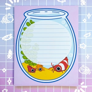 May include: A pink notepad with a blue and white illustration of a fishbowl with three red and white shrimp inside. The fishbowl is filled with blue water and has green plants inside. The notepad has lined paper for writing.