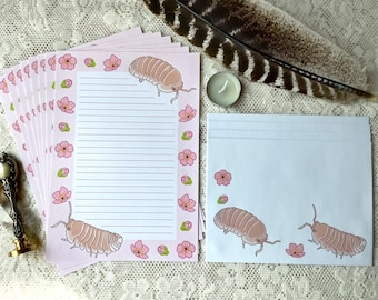 Puede incluir: Juego de papelería rosa con papel de escribir rayado y sobres a juego. El papel y los sobres están decorados con ilustraciones de cochinillas rosas y flores de cerezo. También hay una pluma y una vela en la imagen.