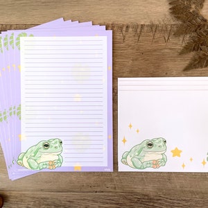 May include: A set of 10 purple stationery note cards with a green frog illustration and gold stars. The cards are lined and have a white border. The cards are packaged with a set of 5 blank white note cards with a green frog illustration, gold stars, and a green leaf.