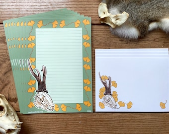 Op de afbeelding: Een set groen en geel briefpapier met een schedel met geweien. Het briefpapier heeft een ginkgo-bladerenrand en gestreept schrijfpapier. Het briefpapier ligt op een houten oppervlak.