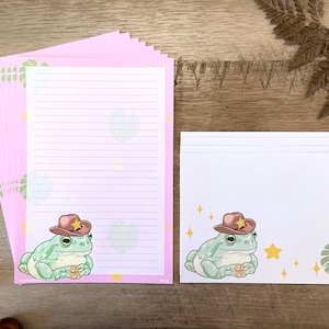 May include: A set of pink and white stationery featuring a cartoon frog wearing a cowboy hat. The frog is surrounded by stars and leaves. The stationery is perfect for writing letters or notes.