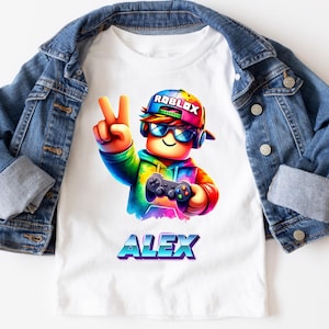 Puede incluir: Camiseta blanca con un personaje colorido de Roblox sosteniendo un mando de videojuegos y haciendo el signo de la paz. El personaje lleva un sombrero arcoíris y gafas de sol. El nombre "ALEX" está impreso en una fuente vibrante y multicolor debajo. Una chaqueta vaquera está sobre la camiseta.