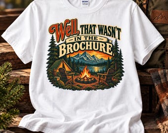 May include: A white short-sleeve t-shirt featuring a graphic print of a campfire scene with a tent, two chairs, a lake, and mountains. The text "WELL, THAT WASN'T IN THE BROCHURE" is printed in a stylized font above the camping illustration.