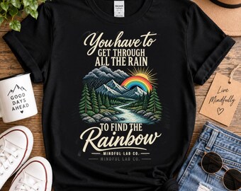 May include: A black short-sleeve t-shirt featuring a graphic of mountains, a river, pine trees, and a rainbow under a sun. The text reads: You have to GET THROUGH ALL THE RAIN TO FIND THE Rainbow MINDFUL LAB CO. The shirt has a crew neck and a tag.